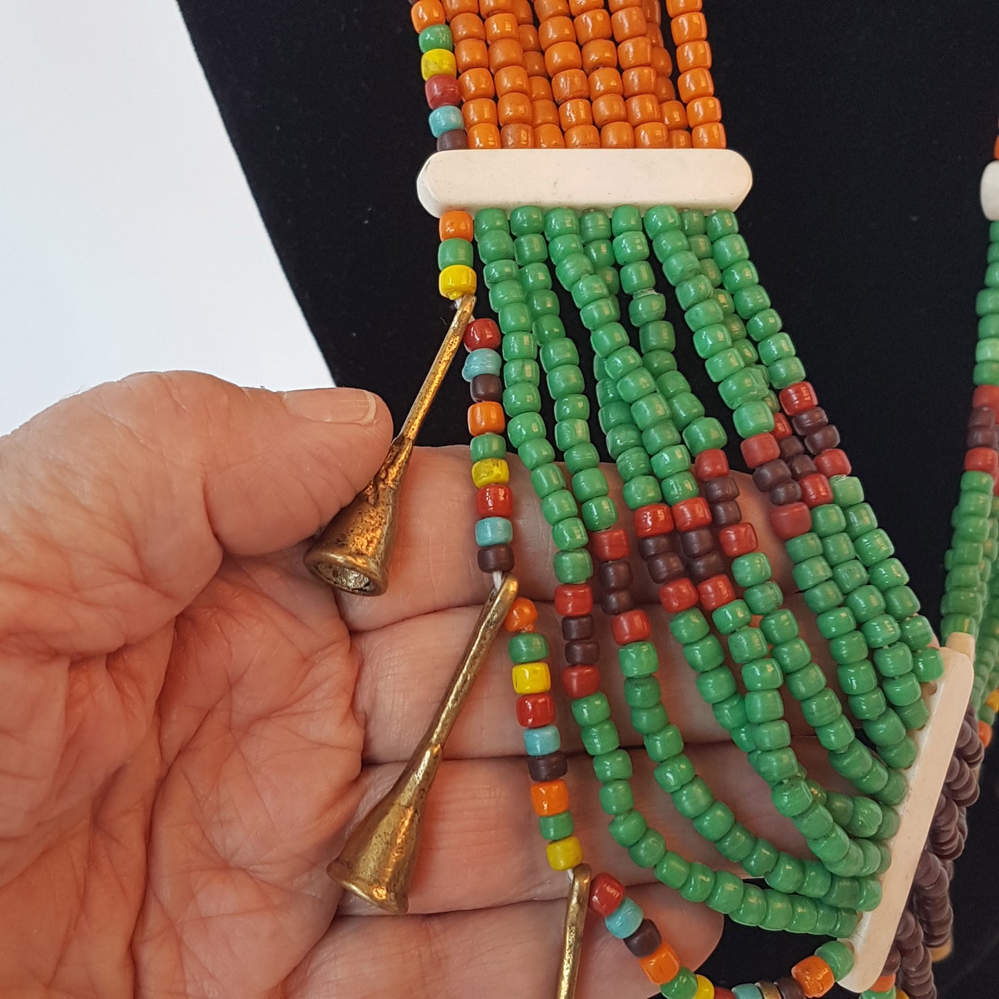 Collier de perles tribal dans une superbe combinaison d'orange, de marron, de vert et de rouille. Fermeture par bouton en os. Détail de corne en métal bronze antique.
