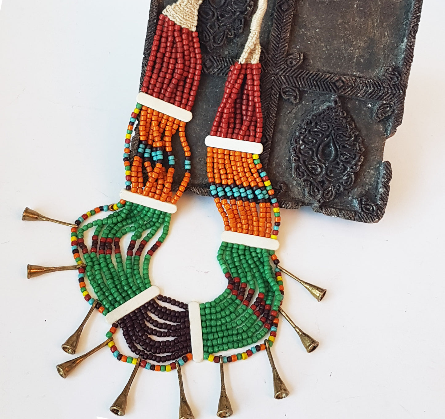 Collier de perles tribal dans une superbe combinaison d'orange, de marron, de vert et de rouille. Fermeture par bouton en os. Détail de corne en métal bronze antique.