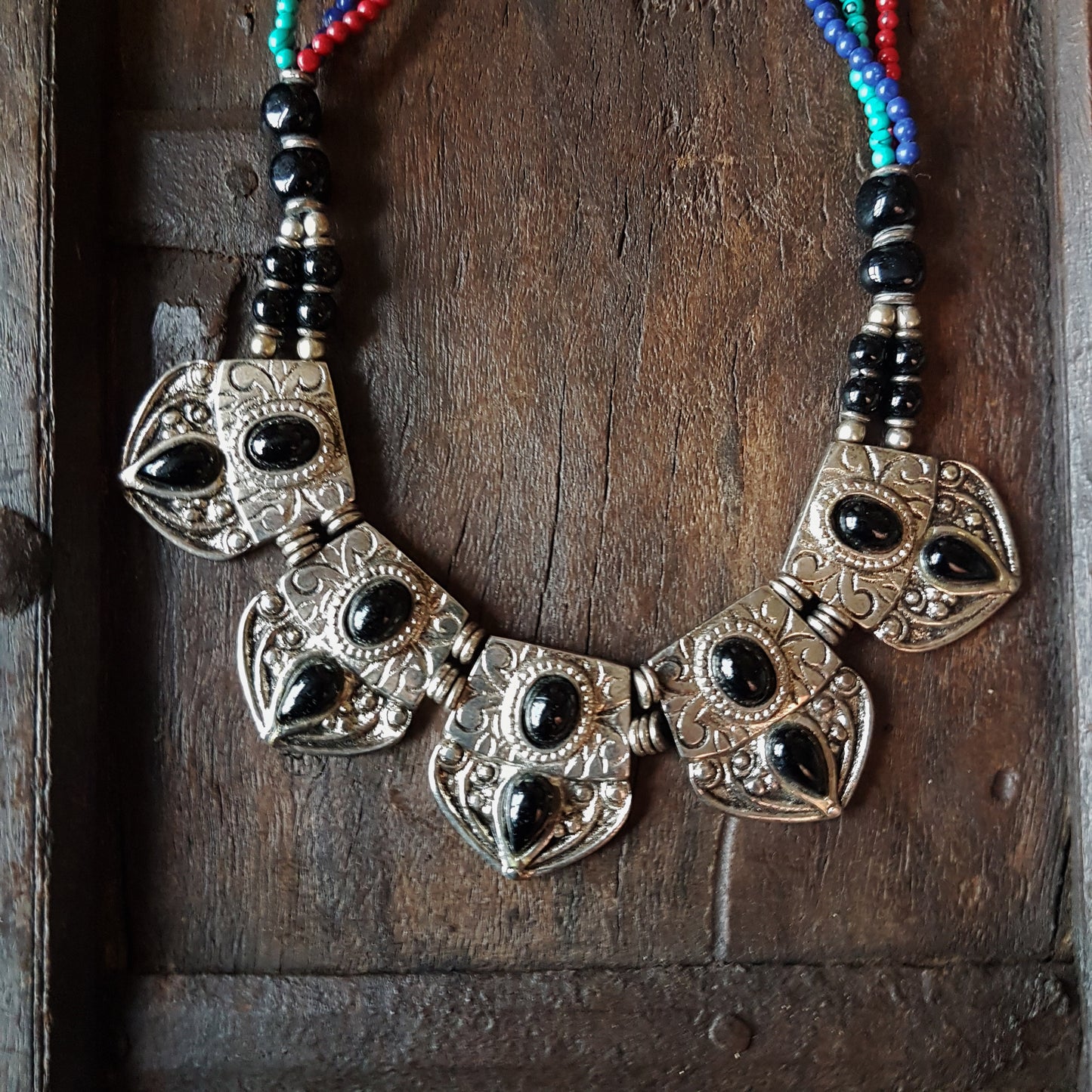 Collier indo-tibétain dramatique avec des pierres d'onyx noires incrustées sur du métal argenté gaufré. Finition corde perlée multicolore.